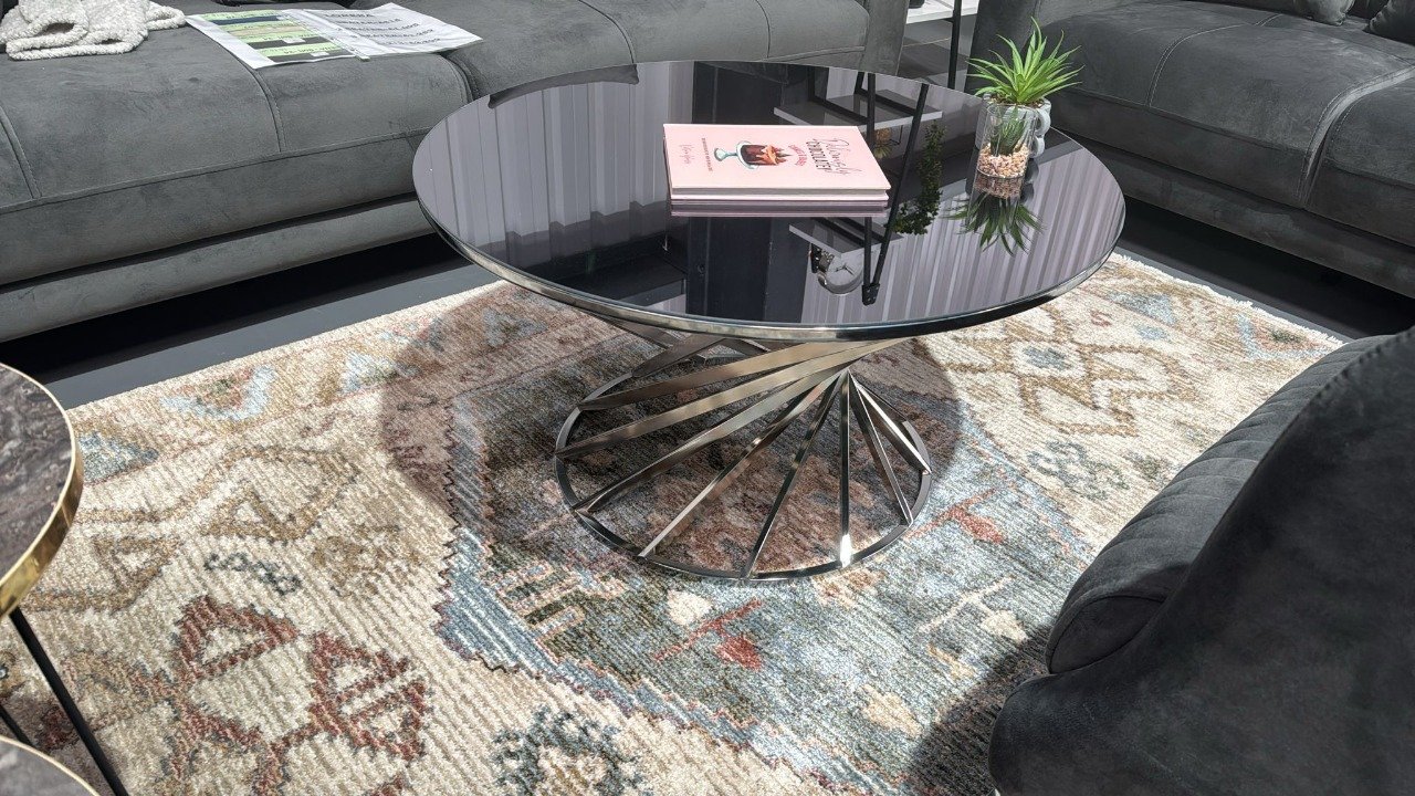 A round glass Arbor Coffee Table with a spiral metal base, surrounded by gray sofas and a patterned rug.