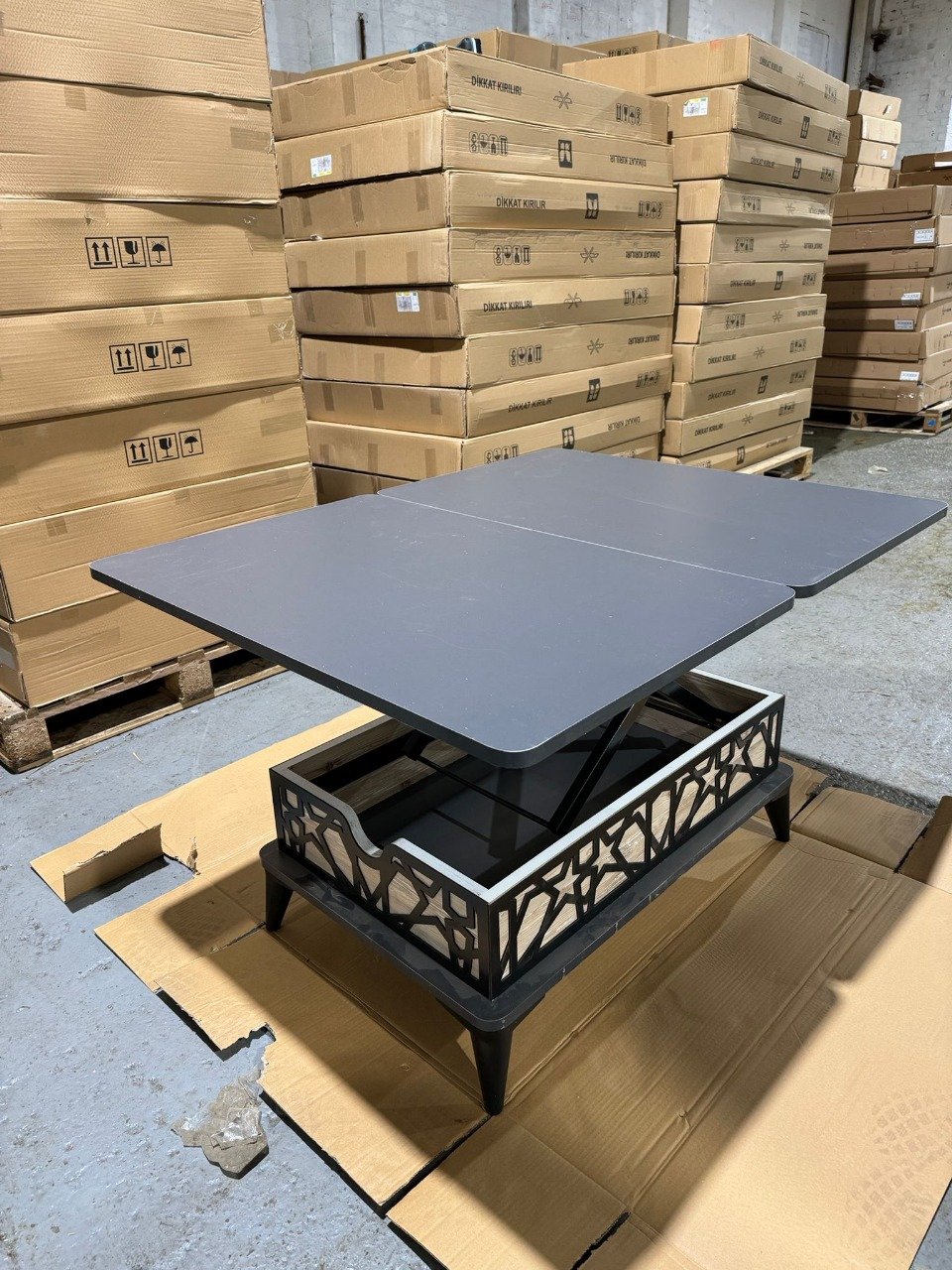A lift-top coffee table in front of stacked cardboard boxes in a warehouse.