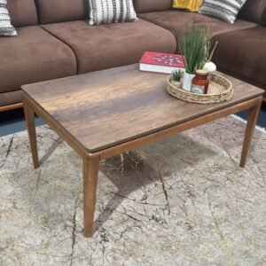 Cleo Coffee Table with a wooden finish and tapered legs, placed on a patterned rug in front of a brown sectional sofa.