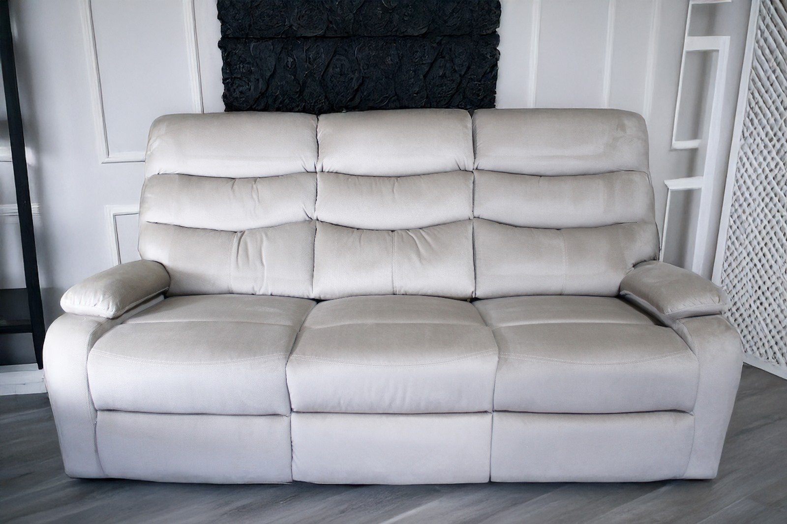 Light gray three-seater sofa with cushioned backrest and armrests against a white paneled wall with black artwork.