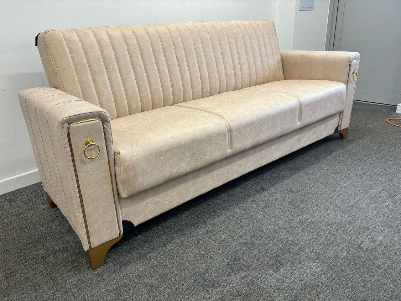 A beige Lucca Sofa Bed with vertical stitching and gold accents against a light-colored wall.