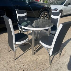 A round glass-top table with four black and white chairs on a concrete surface, with parked cars and a vine-covered fence in the background.