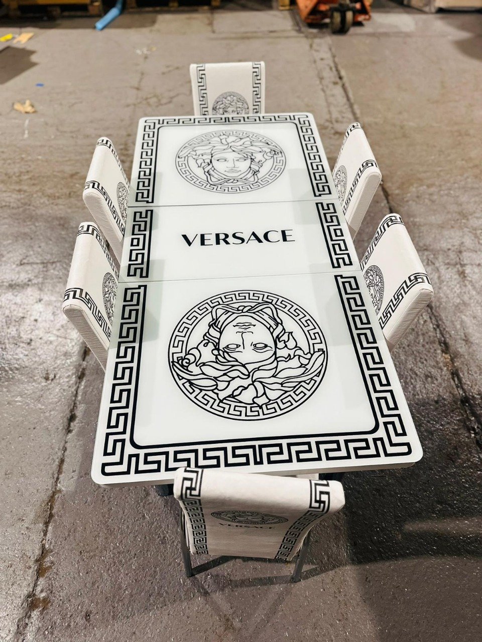 A rectangular dining table set with Greek key designs and Medusa motifs, labeled "VERSACE," on a concrete floor.