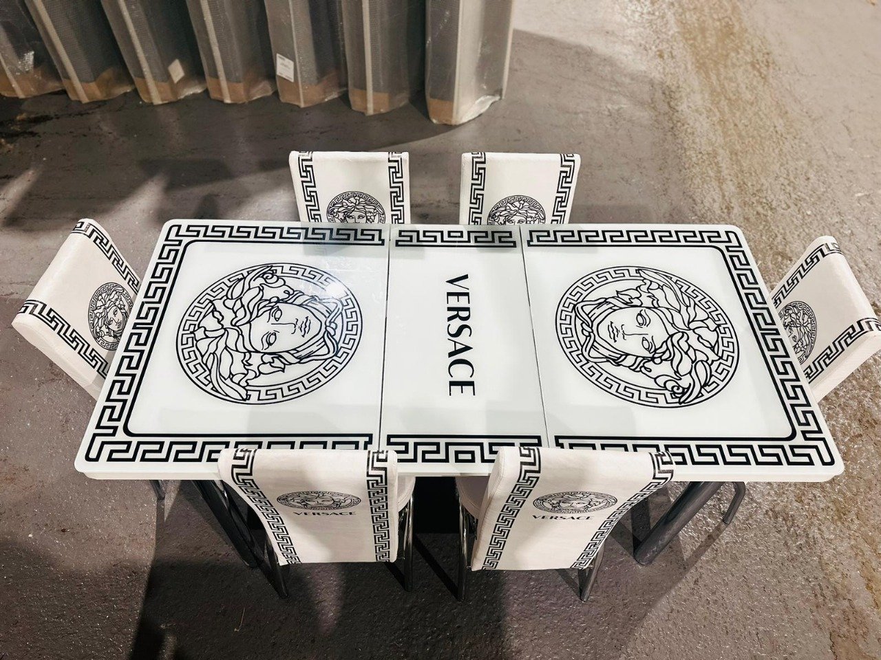 A rectangular dining table set with Greek key designs and Medusa motifs, labeled "VERSACE," on a concrete floor.