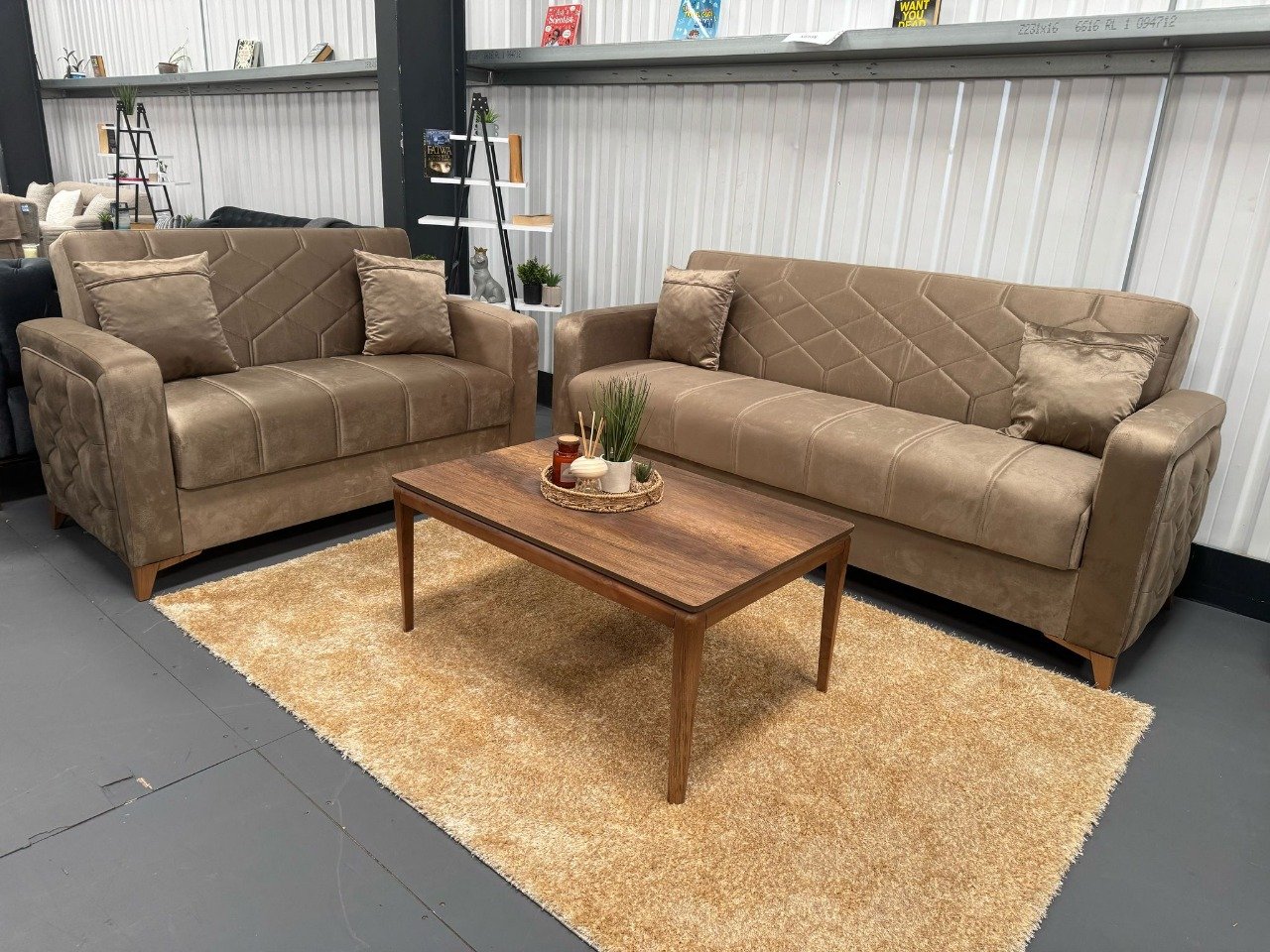 Two Romano Sofa Beds in light brown fabric with a wooden coffee table on a beige rug in a showroom.
