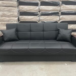 Black leather sofa with wood-accented armrests and two pillows, set against a backdrop of stacked, plastic-wrapped couches.