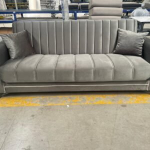A modern grey sofa with vertical stitching and two matching pillows in a warehouse setting.